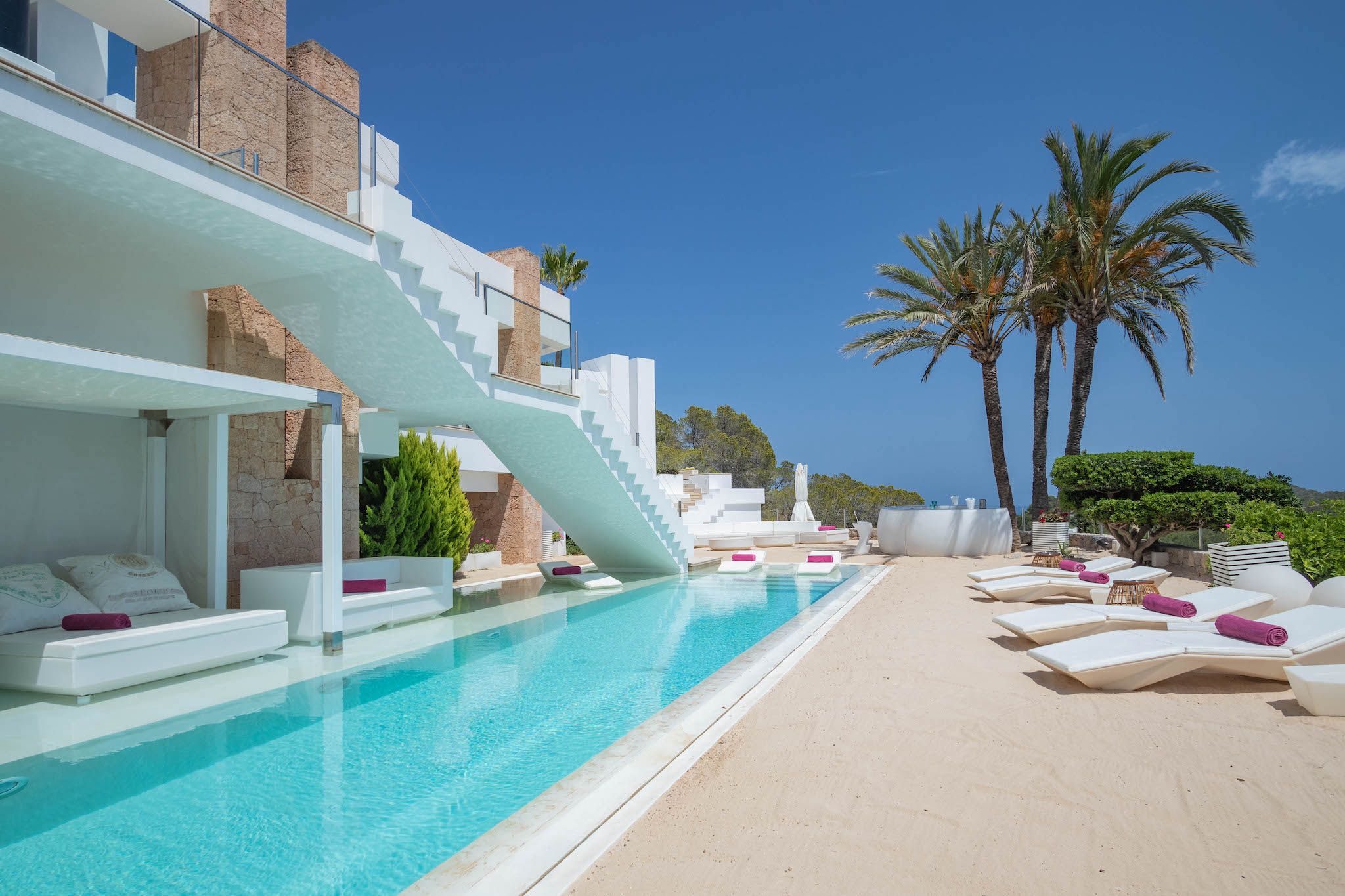 Zona de piscina con una piscina azul cristalina, sillas de descanso blancas y una escalera que conduce a un nivel superior.