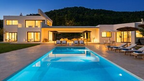Ein modernes, zweistöckiges Haus mit Swimmingpool, Sitzgelegenheiten im Freien und Blick auf einen bewaldeten Hügel.
