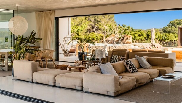 Ein modernes Wohnzimmer mit einer großen Eckcouch, einem Essbereich mit Rattanmöbeln und Blick auf einen Weinberg sowie Sitzgelegenheiten im Freien.