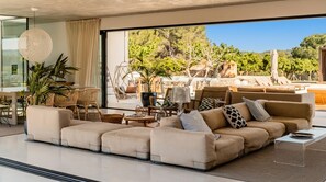 Un salon moderne doté d’un grand canapé d’angle, d’un coin repas avec des meubles en osier et offrant une vue sur un vignoble et un espace de détente extérieur.