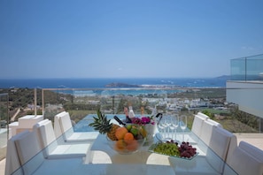A glass table with a fruit bowl, wine glasses, and bottles, overlooking a coastal landscape.