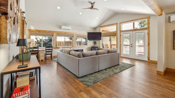 A spacious living area with a gray sofa, wooden flooring, a ceiling fan, and large windows. - Mountain Retreat With Hot Tub, Game Room And Yard 3 Bedroom Home (Cle Elum)