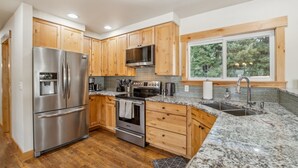 A modern kitchen with wooden cabinets, stainless steel appliances, granite countertops, and a large window with a view of trees. - Mountain Retreat With Hot Tub, Game Room And Yard 3 Bedroom Home (Cle Elum)