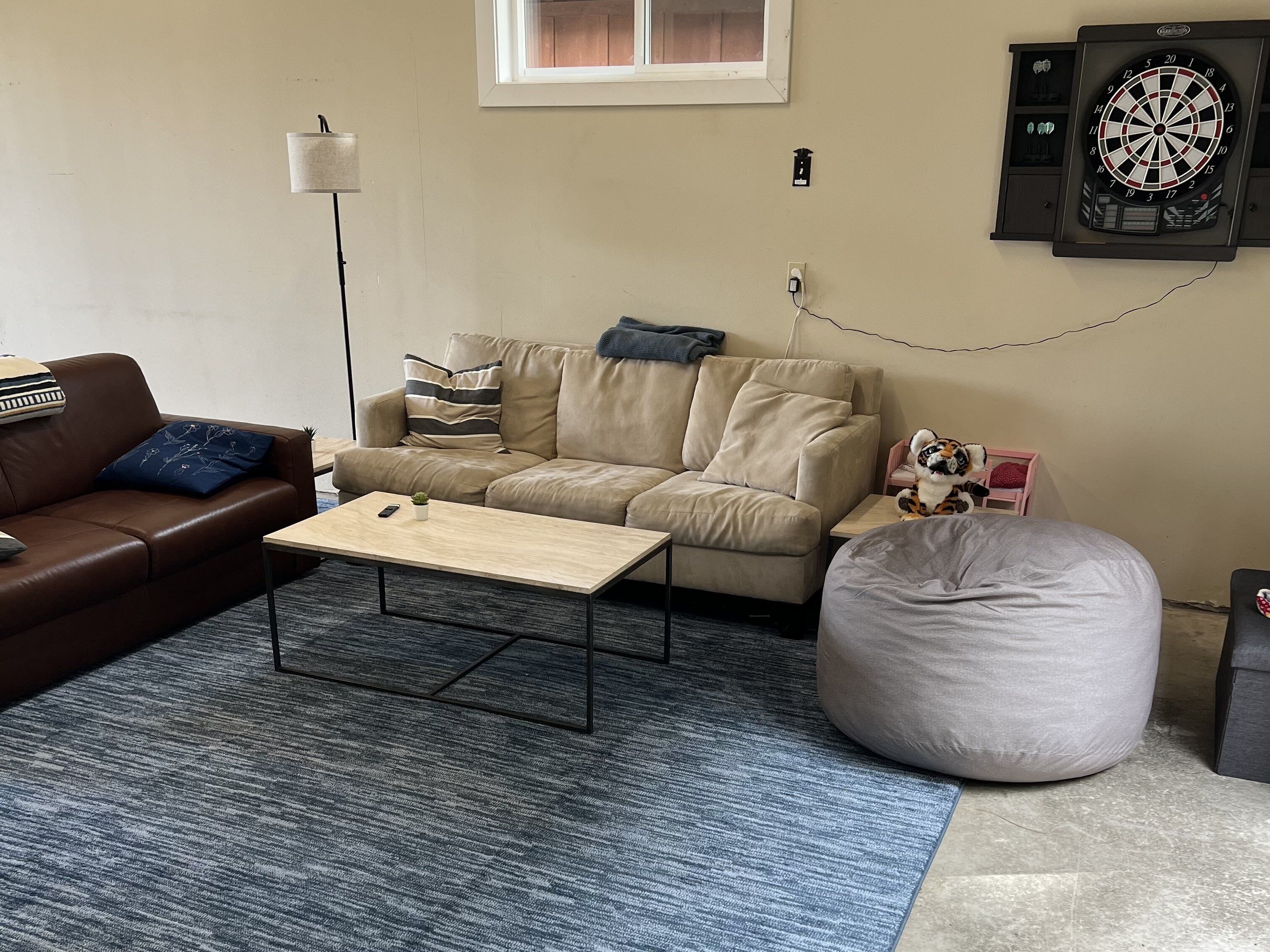Una sala de estar con un sofá de cuero marrón, un sofá beige, una mesa de centro de madera, un sillón de pie gris, un dardo y un estante pequeño con un peluche.