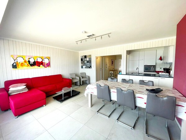A modern living room with a red sofa, a dining table with chairs, and a kitchen area with white cabinets and a microwave.