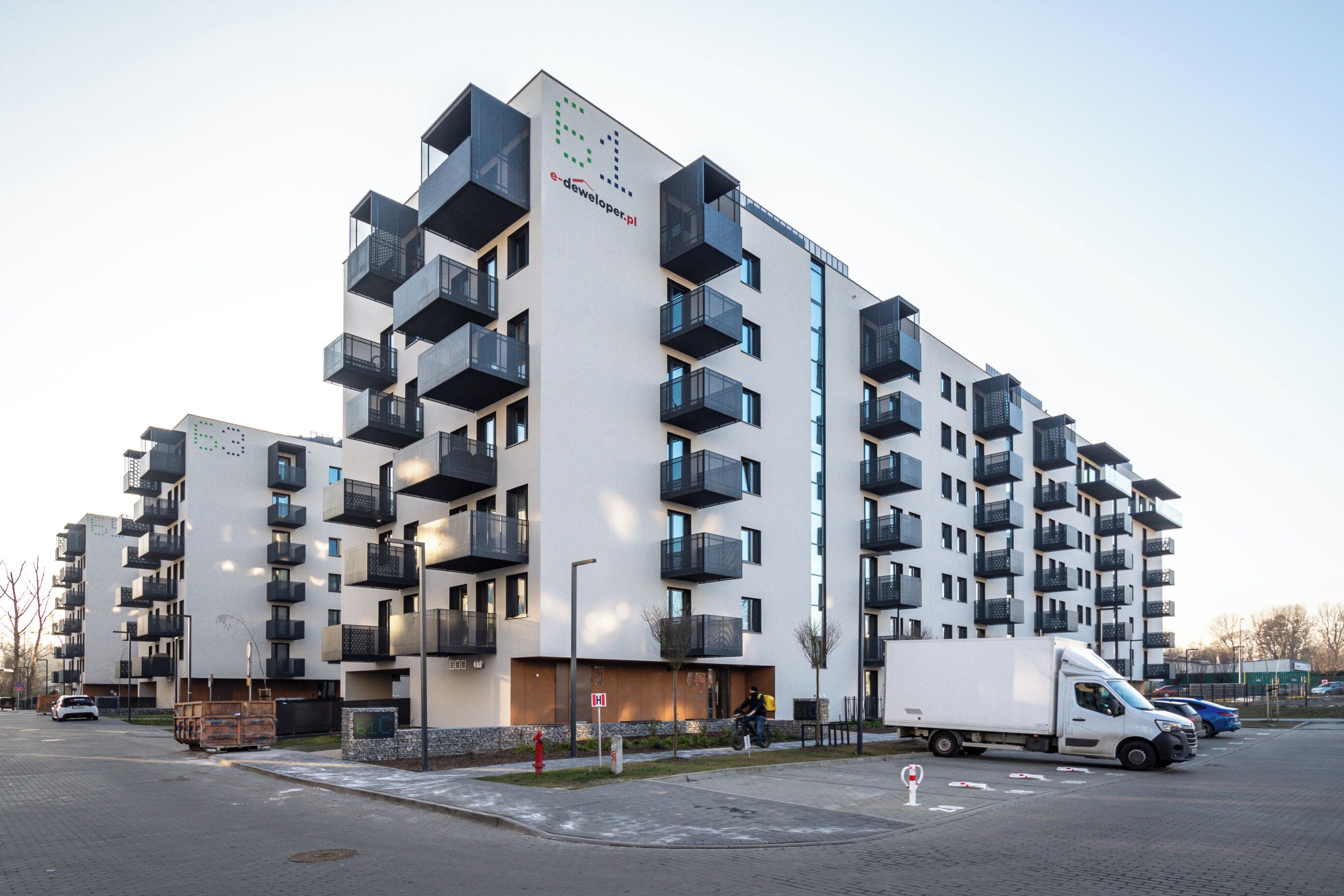 Un immeuble résidentiel moderne à plusieurs étages, doté de balcons, avec un camion garé devant et un ciel dégagé.