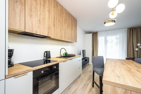 A modern kitchen with wooden cabinets, a black countertop, and a dining area with a wooden table and chairs. - RentPlanet - Apartamenty Ceglana (Katowice)
