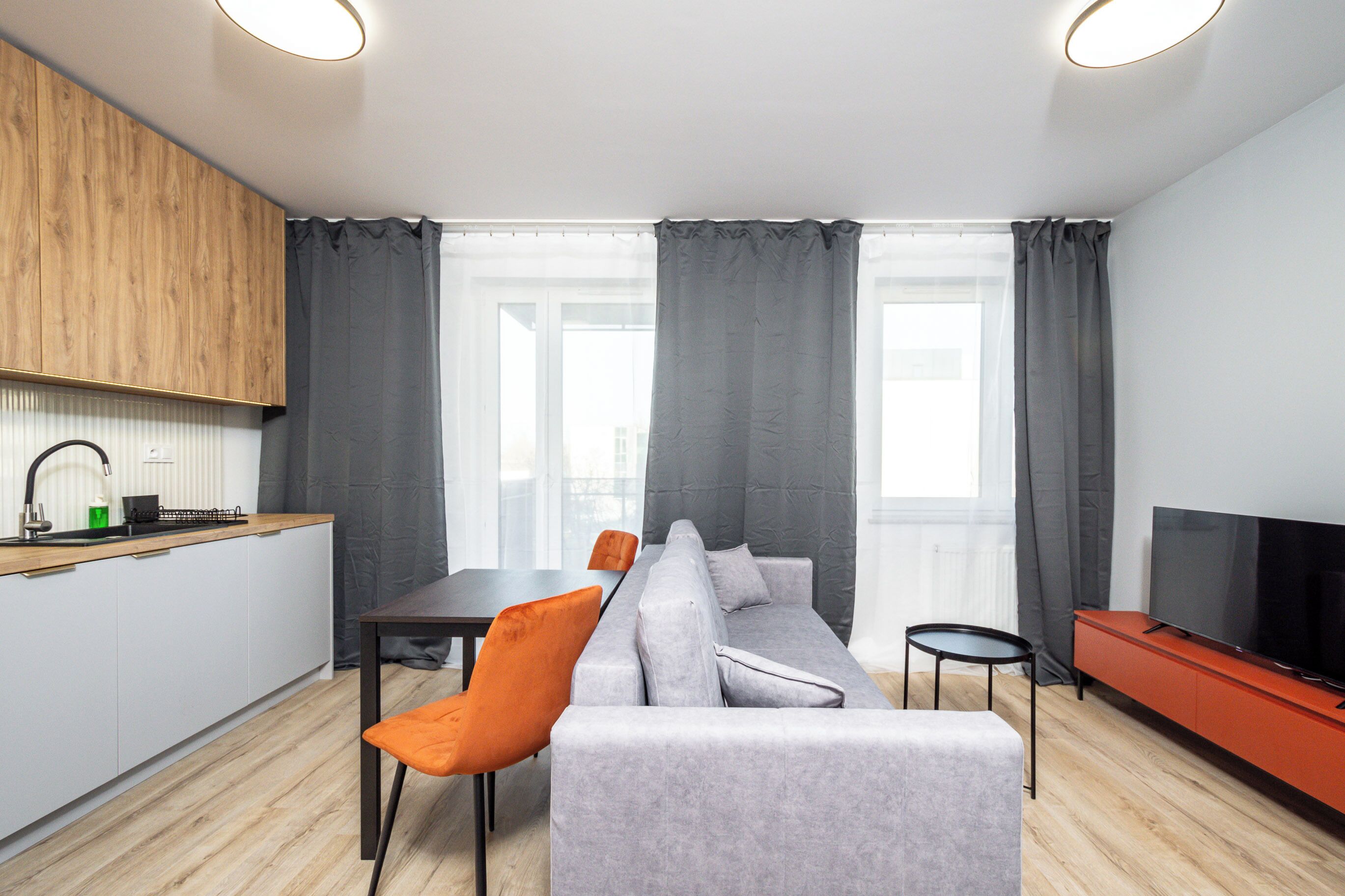 Una sala de estar moderna con un sofá gris, sillas naranjas, una mesita, televisor y una zona de cocina con un armario de madera y un lavamanos.