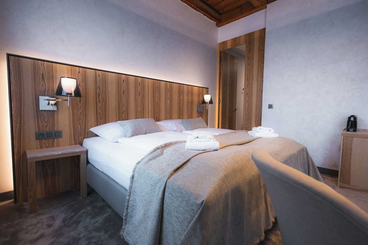 A hotel room with a wooden headboard, two bedside lamps, a bed with a grey blanket, a small bedside table, and a wooden cabinet.