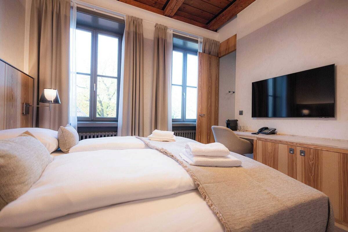A hotel room with a large bed, a desk with a chair, a flat-screen TV, and a window with curtains.