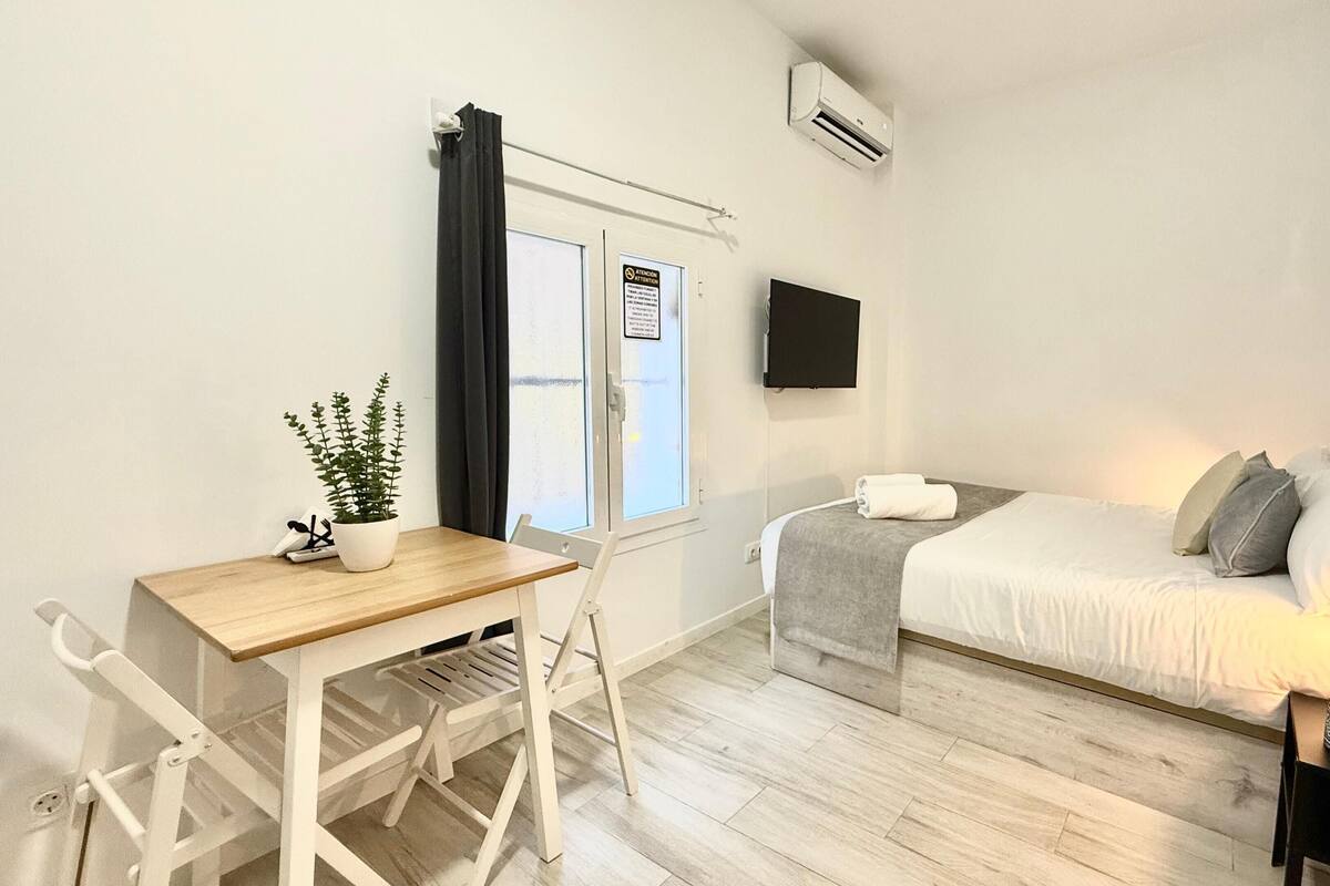 A bedroom with a bed, bedside table, and a desk with a chair. There is a window with curtains, a wall-mounted TV, and a potted plant.