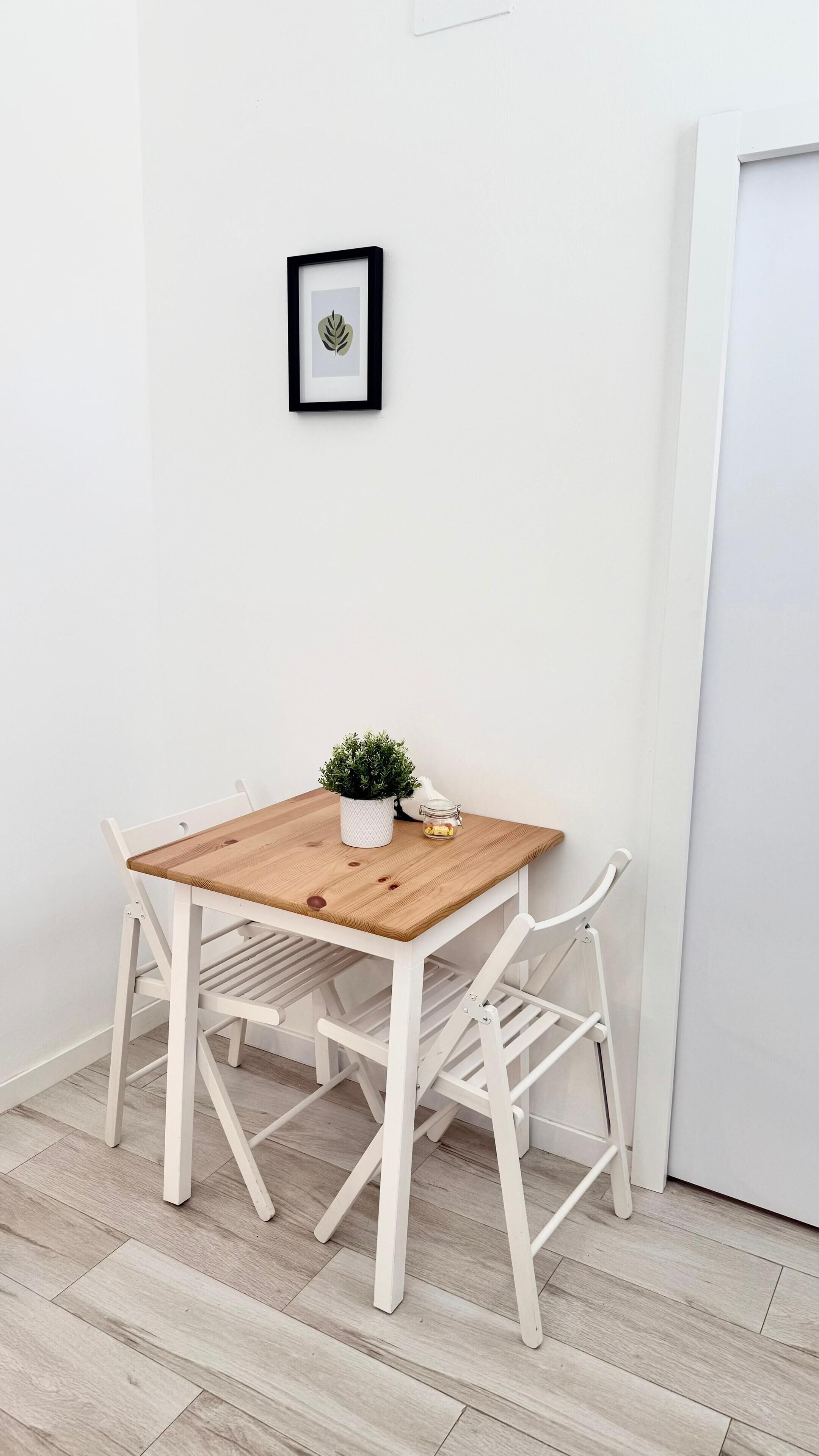 Una pequeña mesa de madera con una planta en maceta y un tarro de vidrio, sobre un fondo de pared blanca con un cuadro colgado arriba.