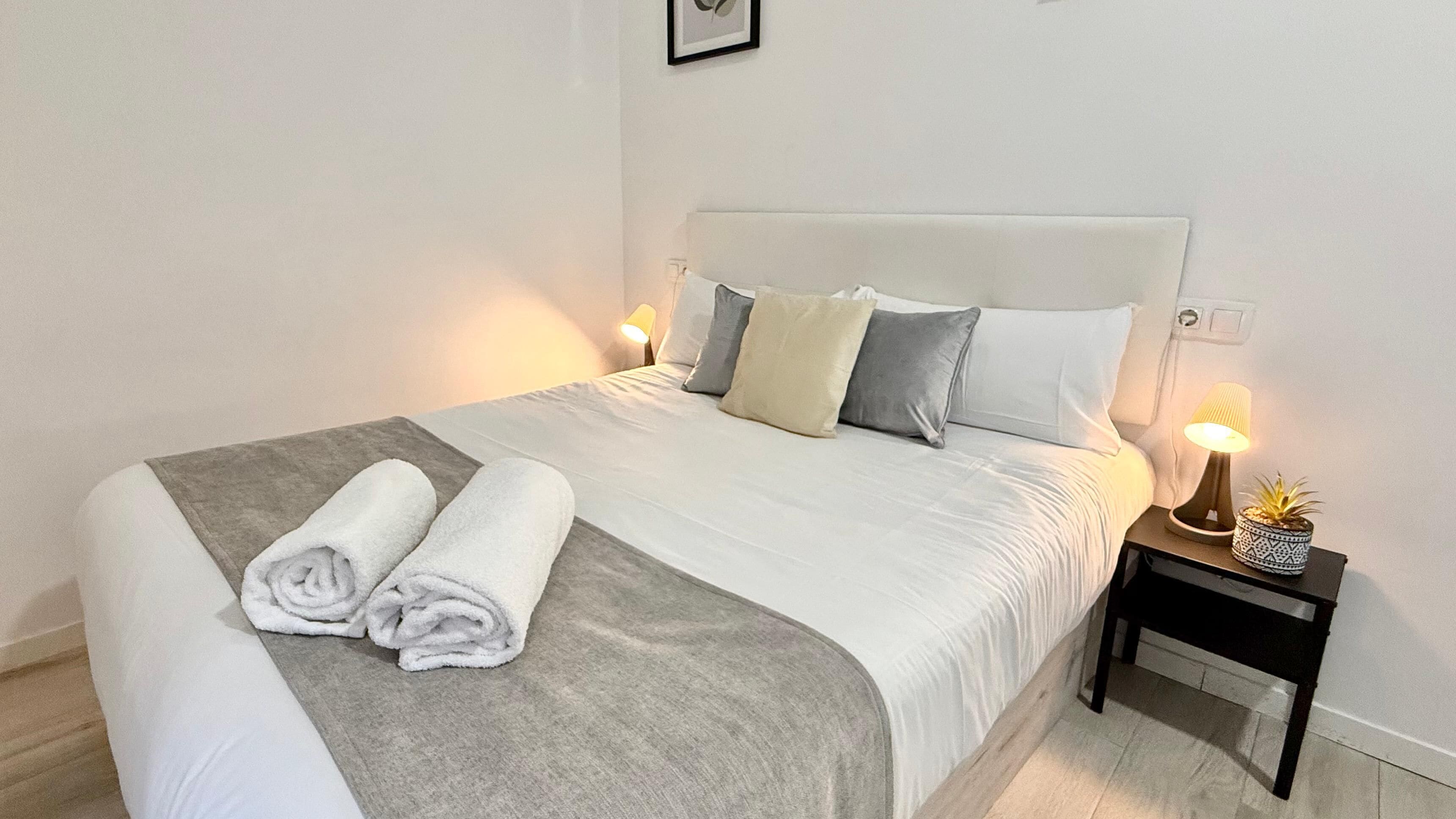 Una cama bien tendida con edredón gris y blanco, dos toallas blancas enrolladas y una manta gris. Hay dos lámparas de noche con pantallas decorativas y una pequeña planta en maceta sobre una mesita de noche de madera junto a la cama.