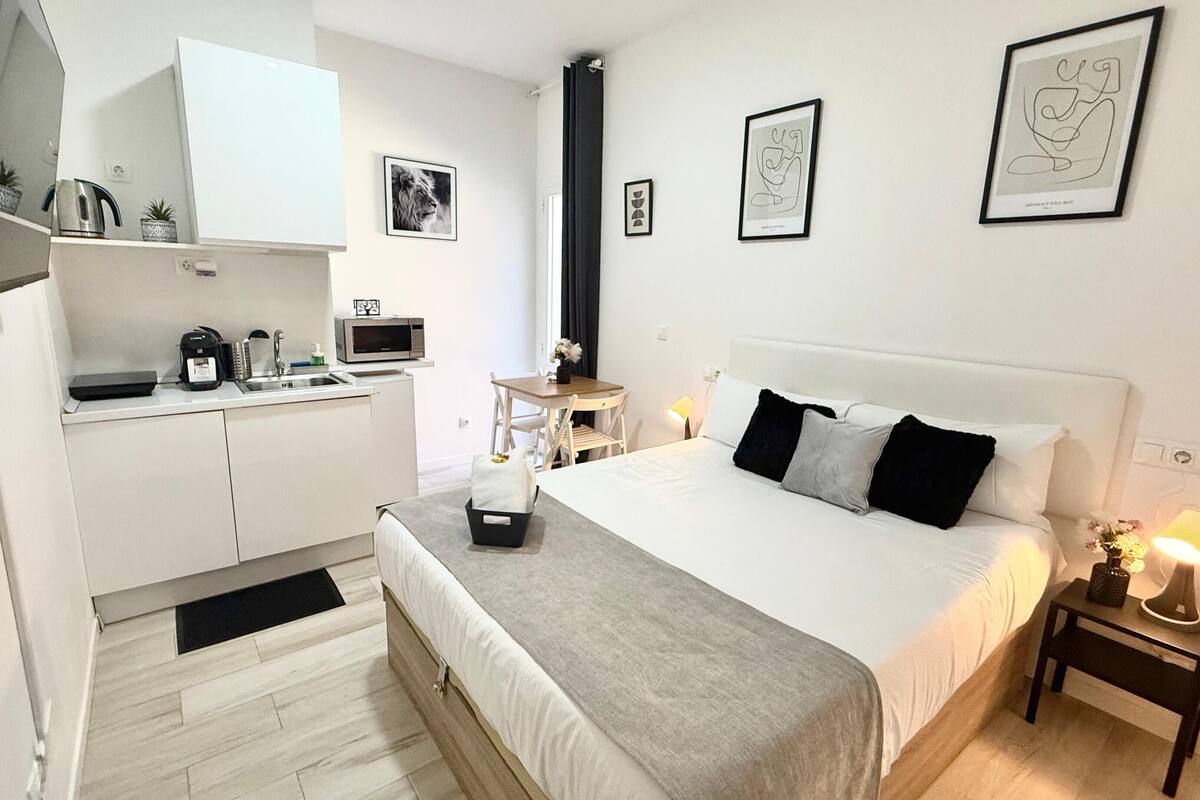 A modern hotel room with a bed, bedside table, lamp, and a kitchenette area with a microwave and kettle.