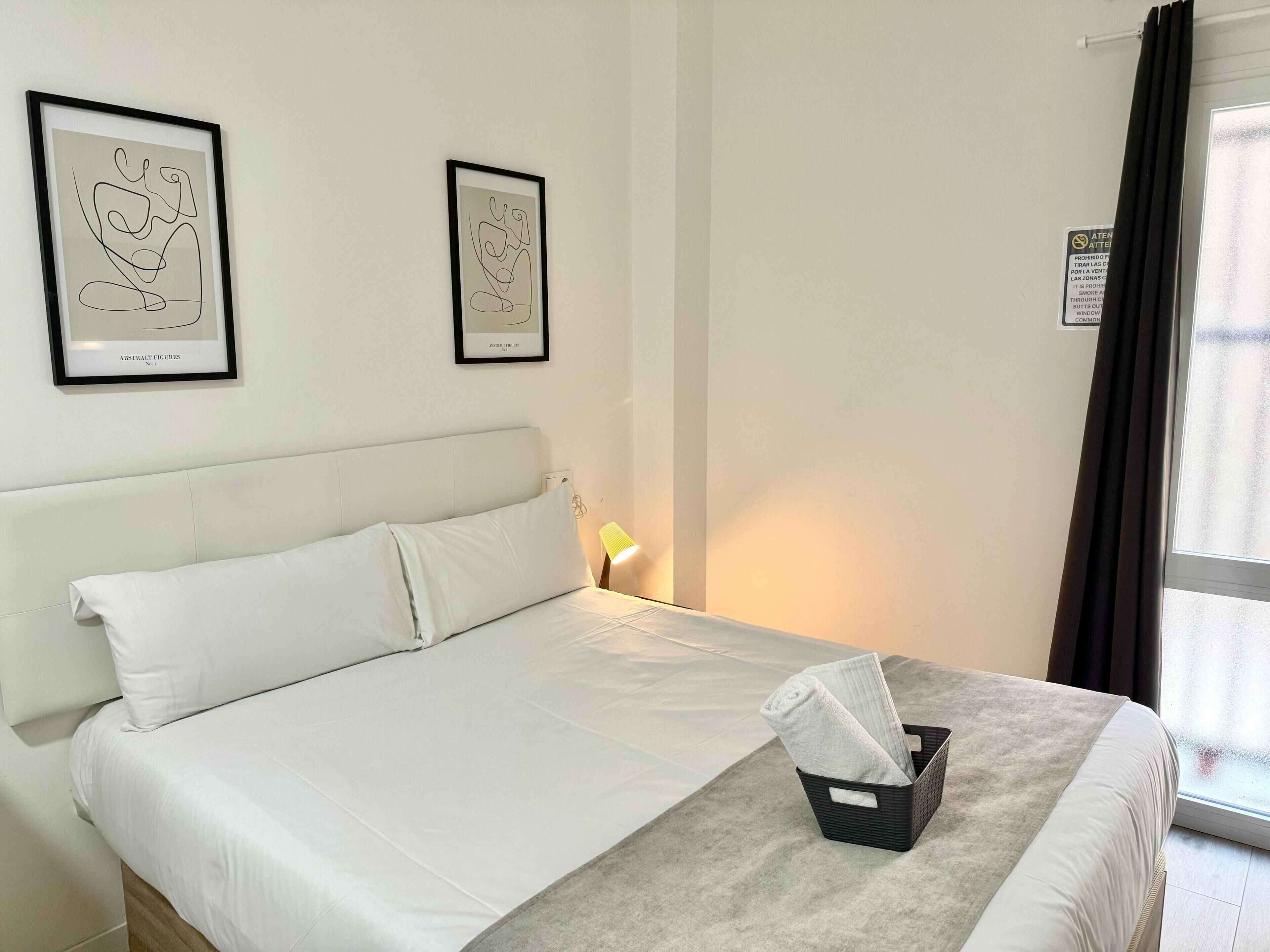 Een net opgemaakt bed met witte lakens, een zwart nachtkastje met een lamp, en twee abstracte kunstwerken aan de muur.