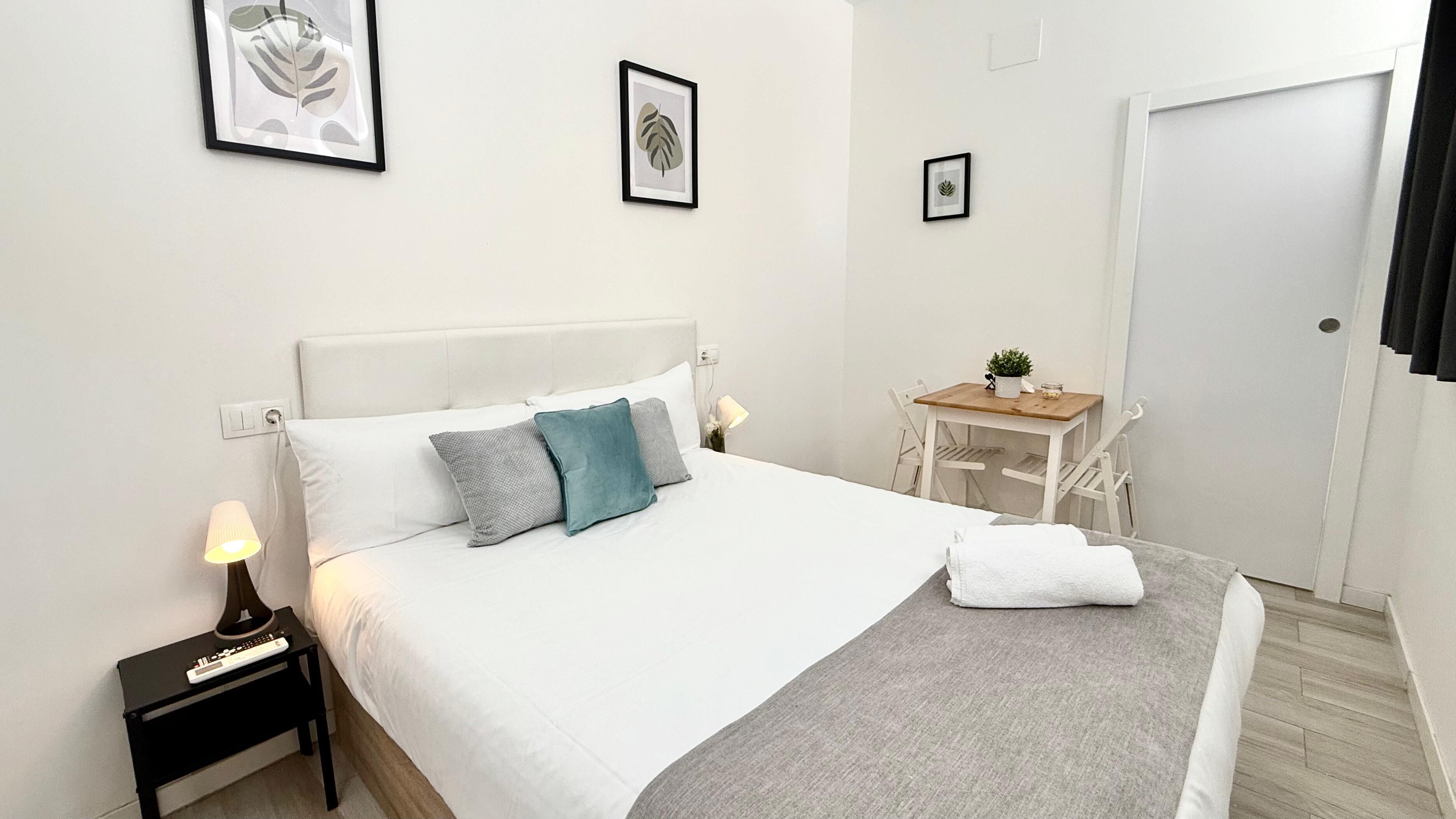 Une chambre d’hôtel bien rangée, avec un lit, une table de chevet, une lampe et une petite table en bois sur laquelle est posée une plante.