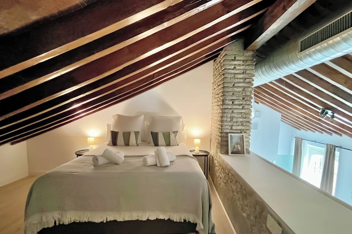 Un dormitorio con techo inclinado, pared de acento de piedra, cama con almohadas, dos lámparas de noche y una ventana con cortinas.