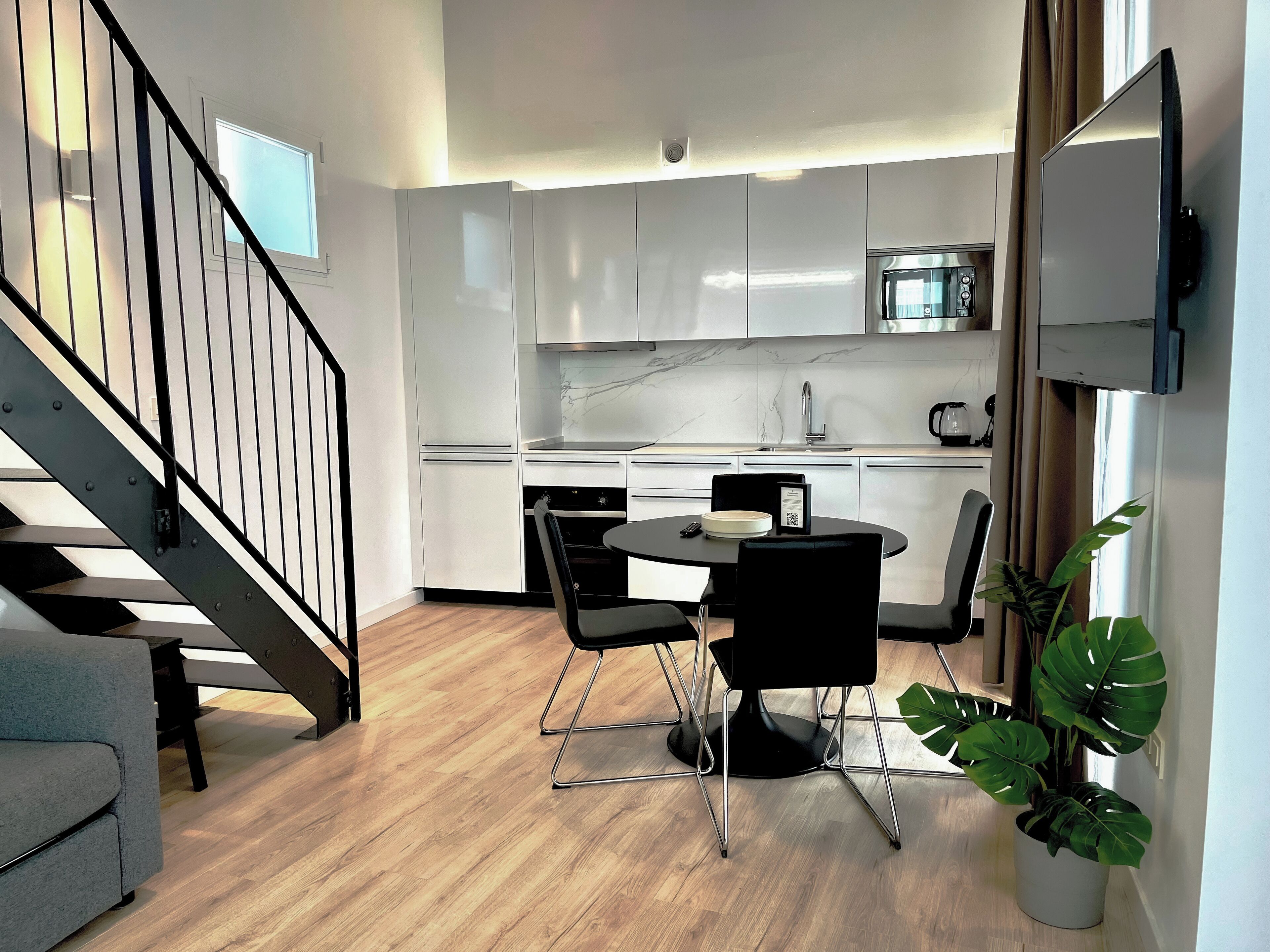 A modern kitchen with white cabinets, a black dining table, and chairs. There is a staircase with black railings leading to another level.