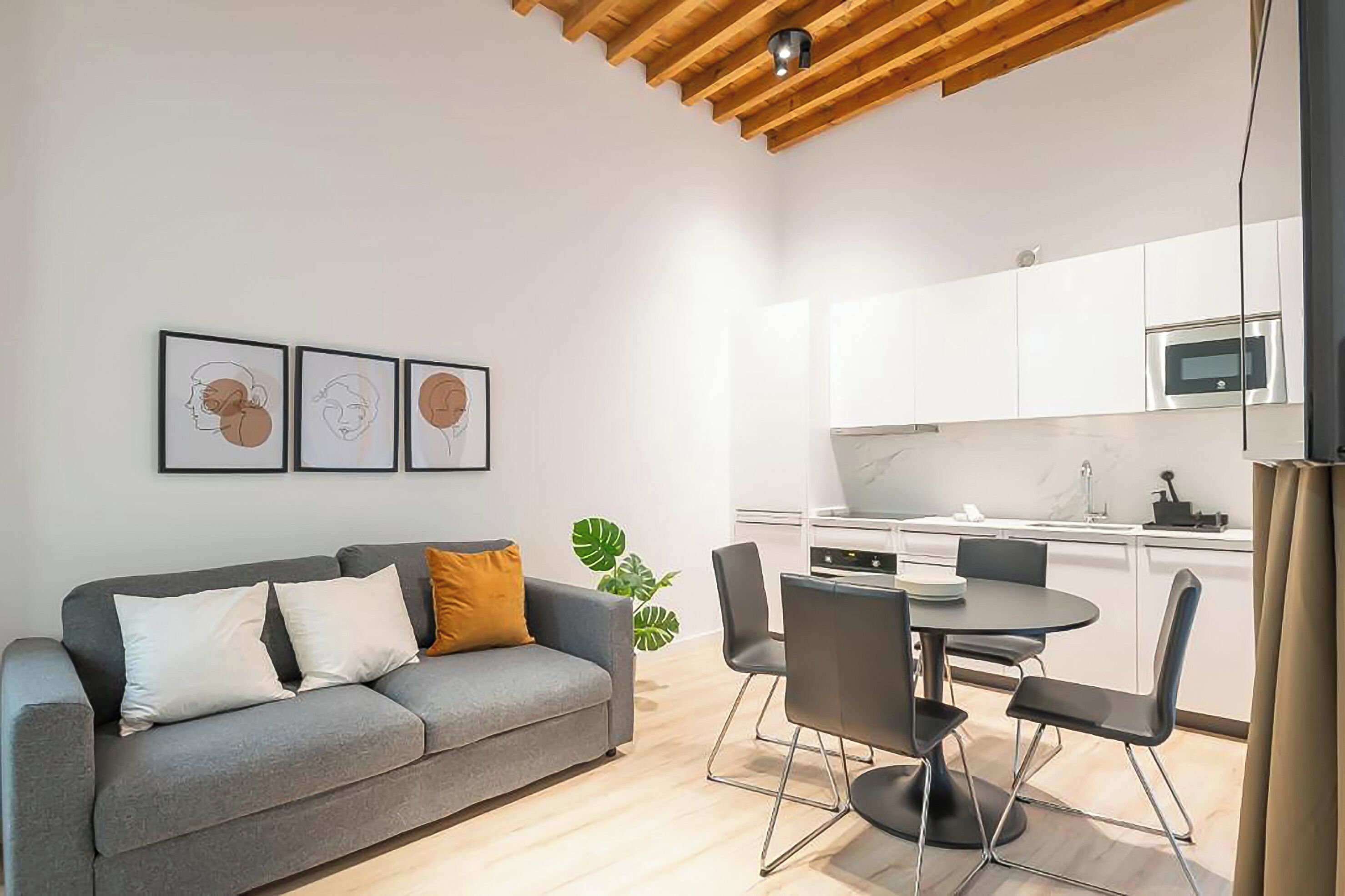 Uma sala de estar moderna com um sofá cinzento, uma mesa de jantar com cadeiras, uma área de cozinha com armários brancos e um teto com vigas de madeira.