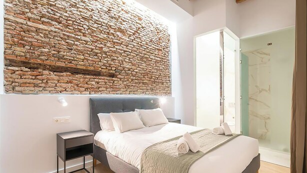 A modern bedroom with a stone wall, a bed with white linens, a bedside table, and a glass shower door. - Feelathome Conde Apartments (Málaga)