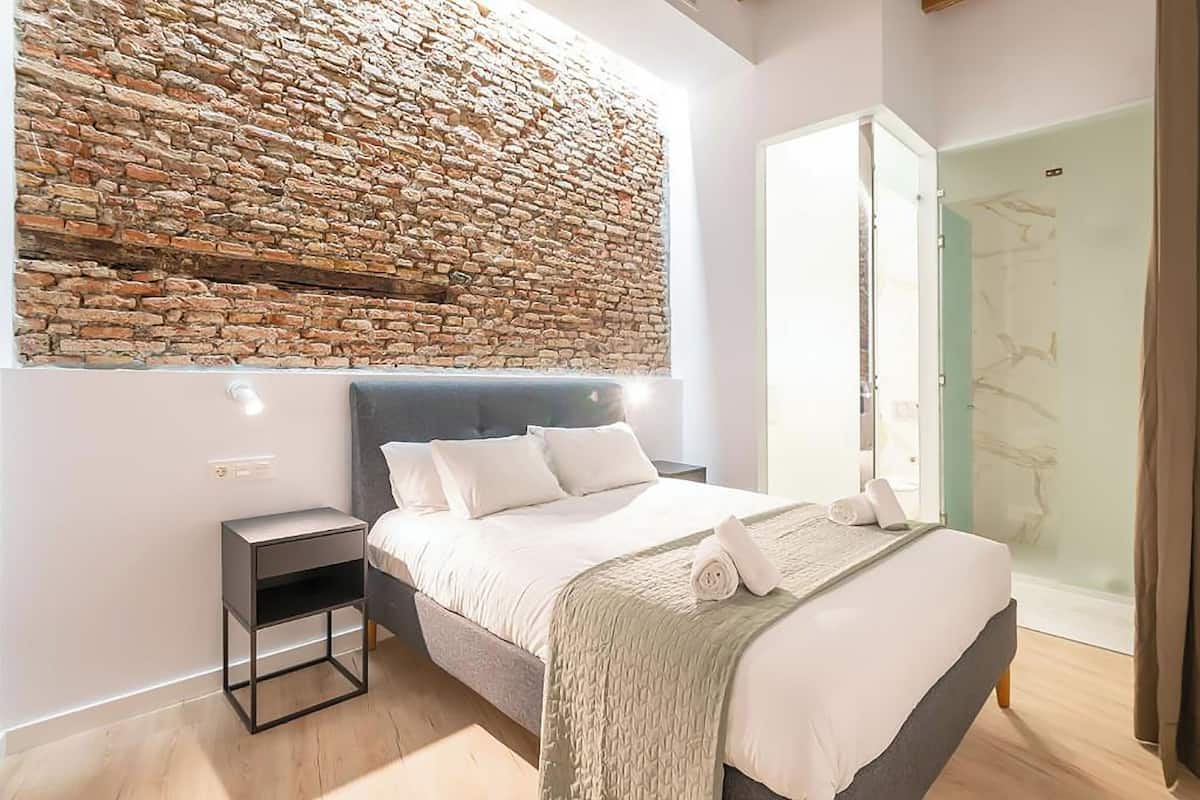 Un dormitorio moderno con pared de piedra, cama con ropa de cama blanca, mesita de noche y puerta de ducha de cristal.