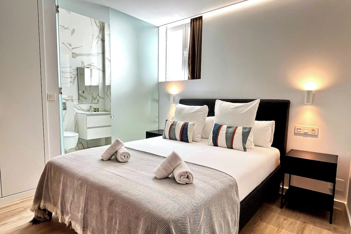Una habitación de hotel moderna con una cama grande, una mesita de noche y un baño visible a través de un panel de vidrio.