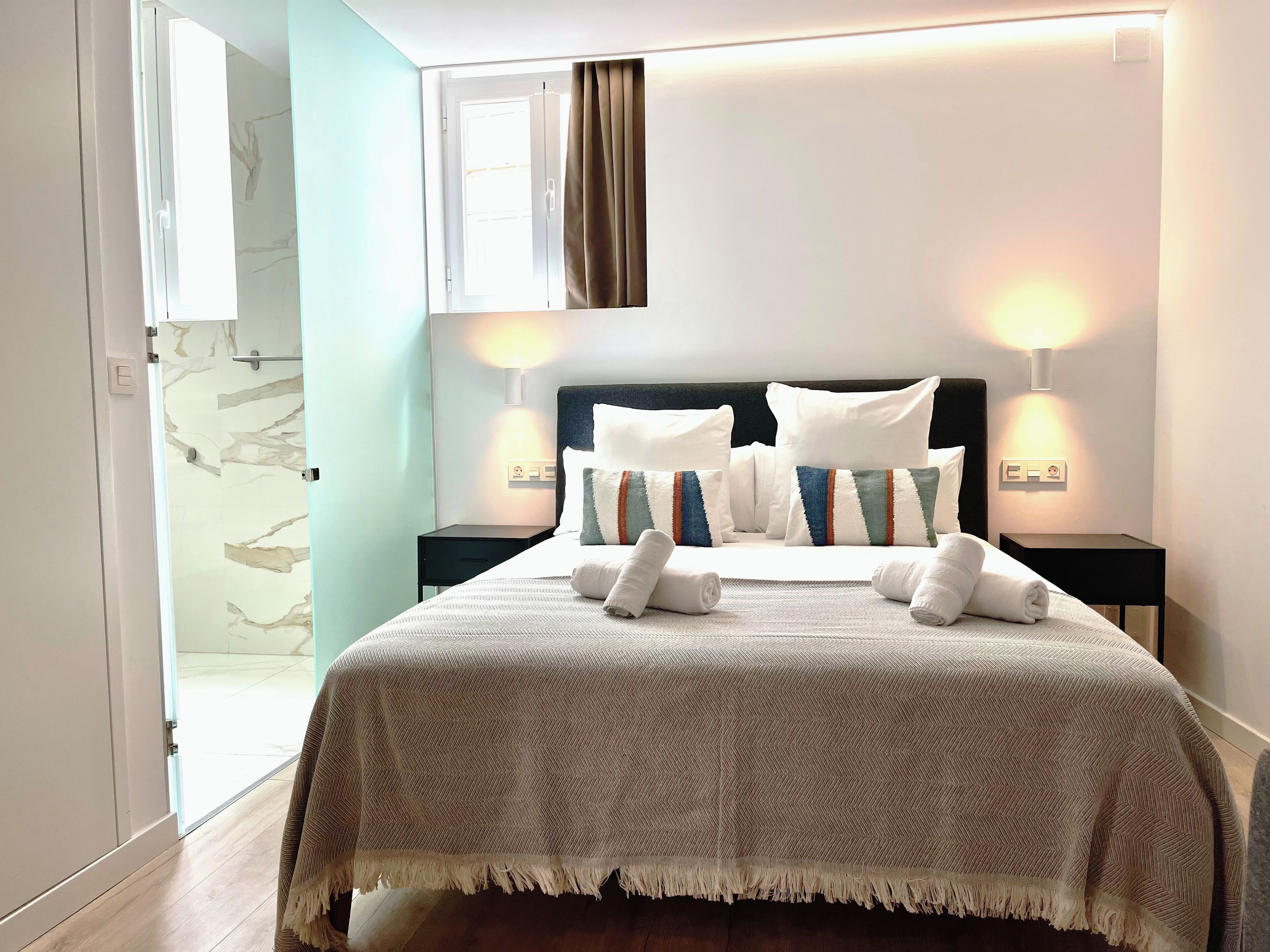 A neatly made bed with a striped pillow, two bedside lamps, and a view of the hallway.