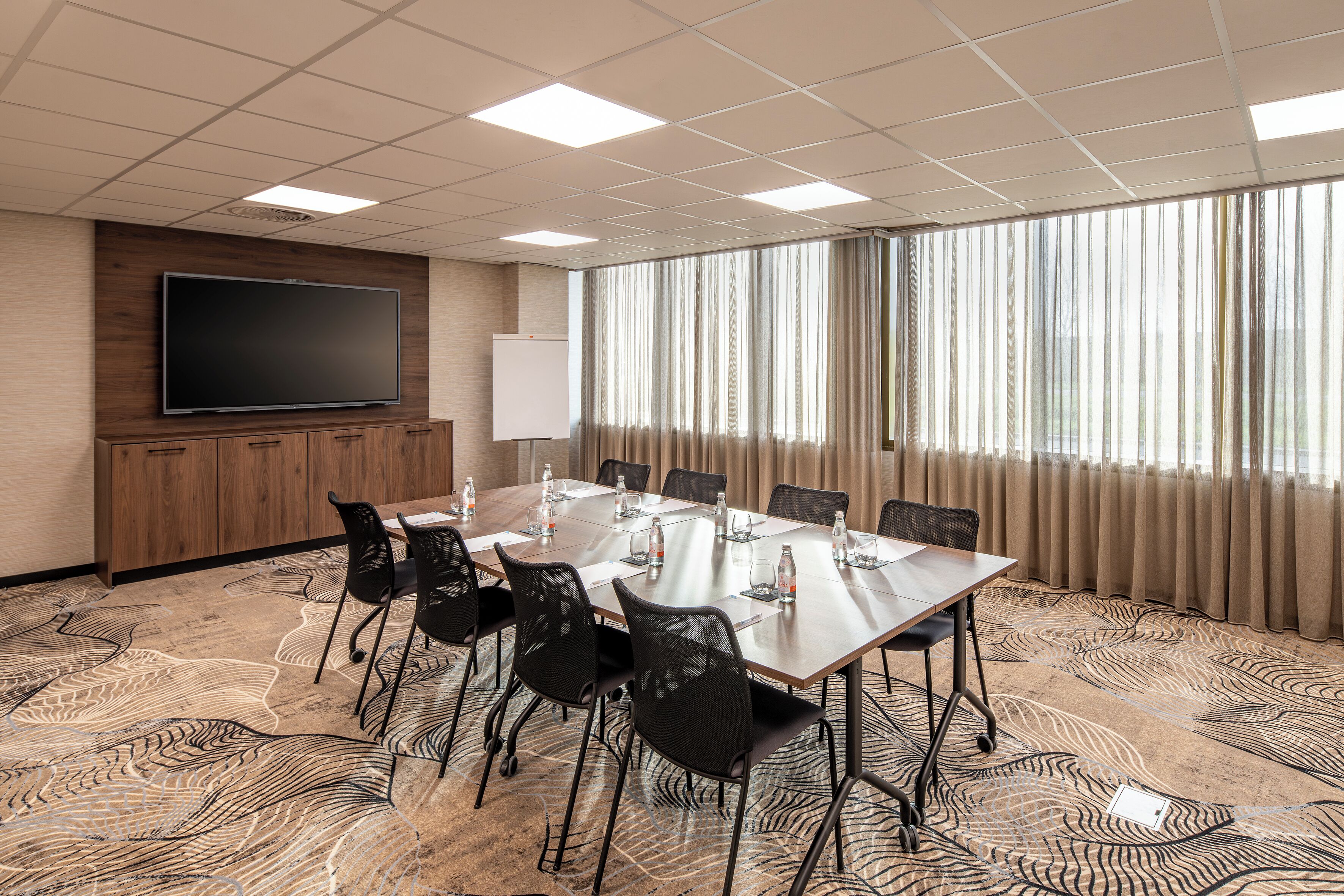 Une salle de conférence équipée d’une longue table, de chaises, d’un téléviseur à écran plat, d’un tableau blanc et de grandes fenêtres avec des rideaux.