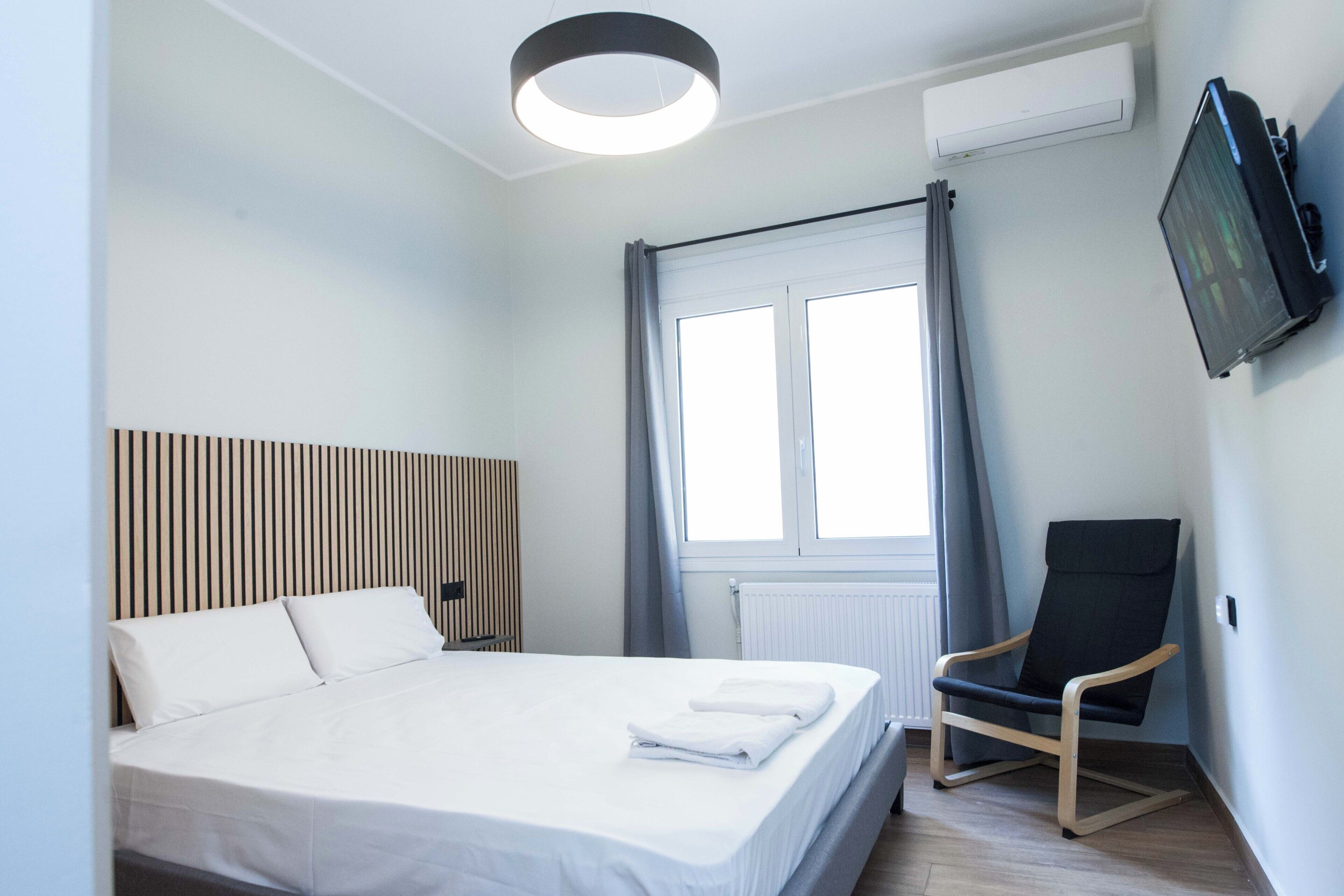 A modern hotel room with a large bed, a flat-screen TV mounted on the wall, a window with curtains, and a wooden chair.