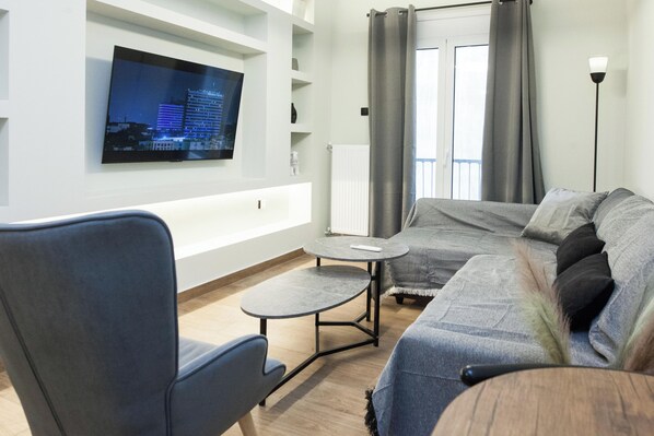 Un salon moderne avec un canapé gris, un fauteuil bleu, une petite table ronde et un téléviseur à écran plat fixé au mur.
