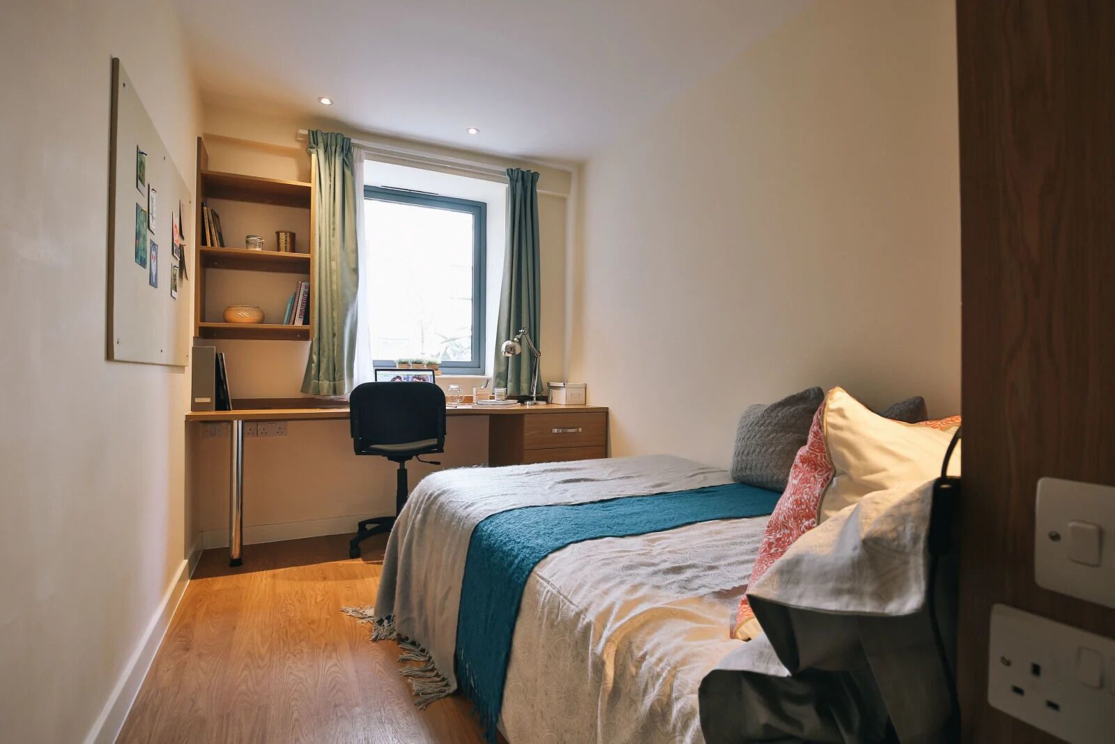 A bedroom with a bed, a desk, and a chair. There is a window with curtains, a shelf with books, and a power outlet.