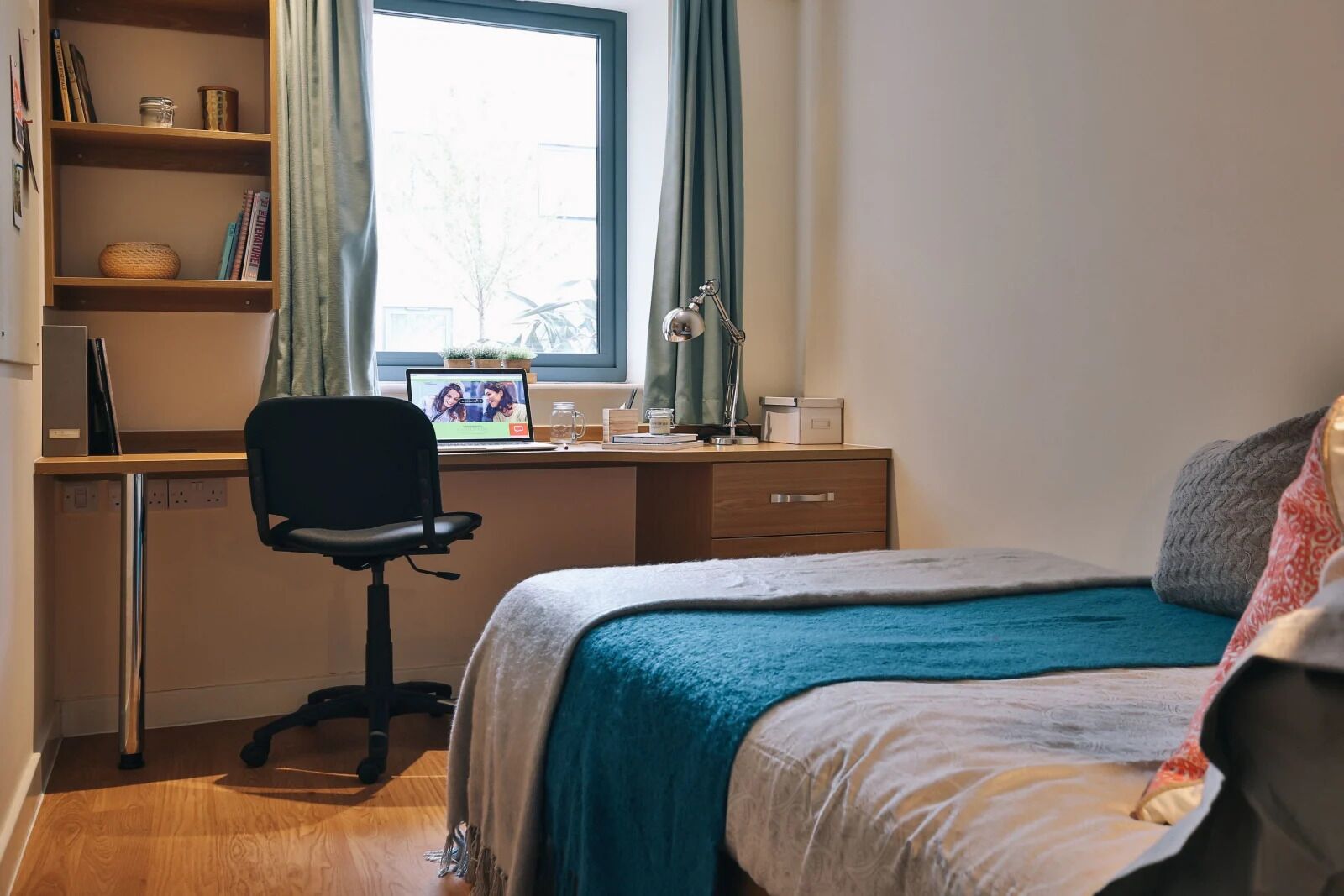 Un dormitorio con una cama, un escritorio con un ordenador portátil, una silla y una ventana con cortinas.