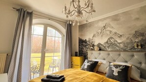 A bedroom with a bed, a chandelier, a mountain mural, and a view of the outdoors.