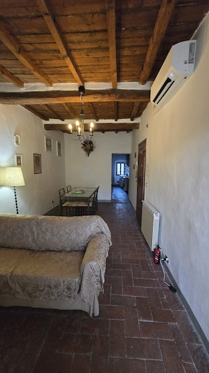 A room with a wooden ceiling, a sofa, a dining table with chairs, a hanging light fixture, and a radiator.