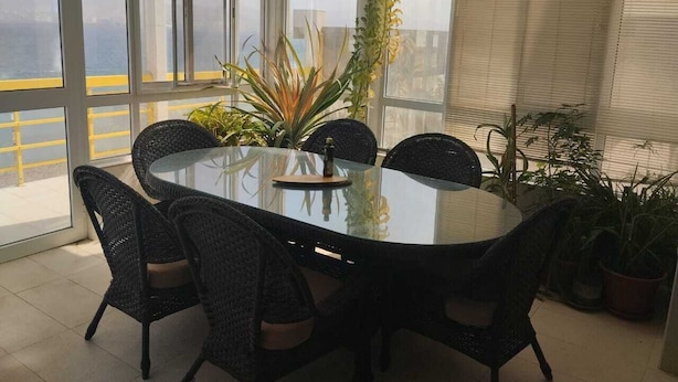 A round glass table with black wicker chairs, surrounded by potted plants, in a bright room with large windows.