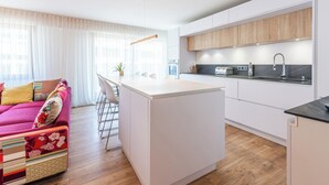 A modern kitchen with white cabinets, a central island, and a dining area with a colorful sofa. - La Quinta - Appartement en Centre ville (Annecy)