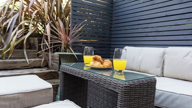 A wicker table with a glass top, a white cushioned bench, and a potted plant in the background.