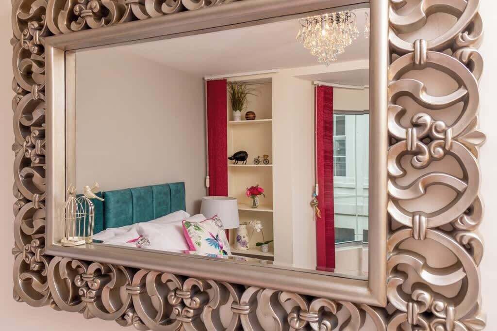 Une chambre à coucher avec un grand miroir décoratif, une tête de lit recouverte d’un tissu turquoise et un lustre.