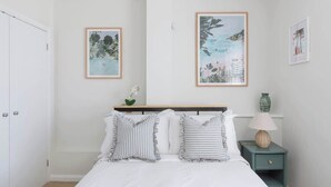 Ein Schlafzimmer mit einem Bett, Kissen, einem Nachttisch, einer Lampe und zwei gerahmten Bildern an der Wand.