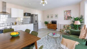 Eine moderne Küche mit weißen Schränken, einem Edelstahl-Kühlschrank und einer Mikrowelle. Ein Esstisch mit Stühlen und eine Schüssel mit Obst sind ebenfalls vorhanden. Das Wohnzimmer verfügt über ein grünes Sofa, einen gemusterten Teppich und einen Fernseher.