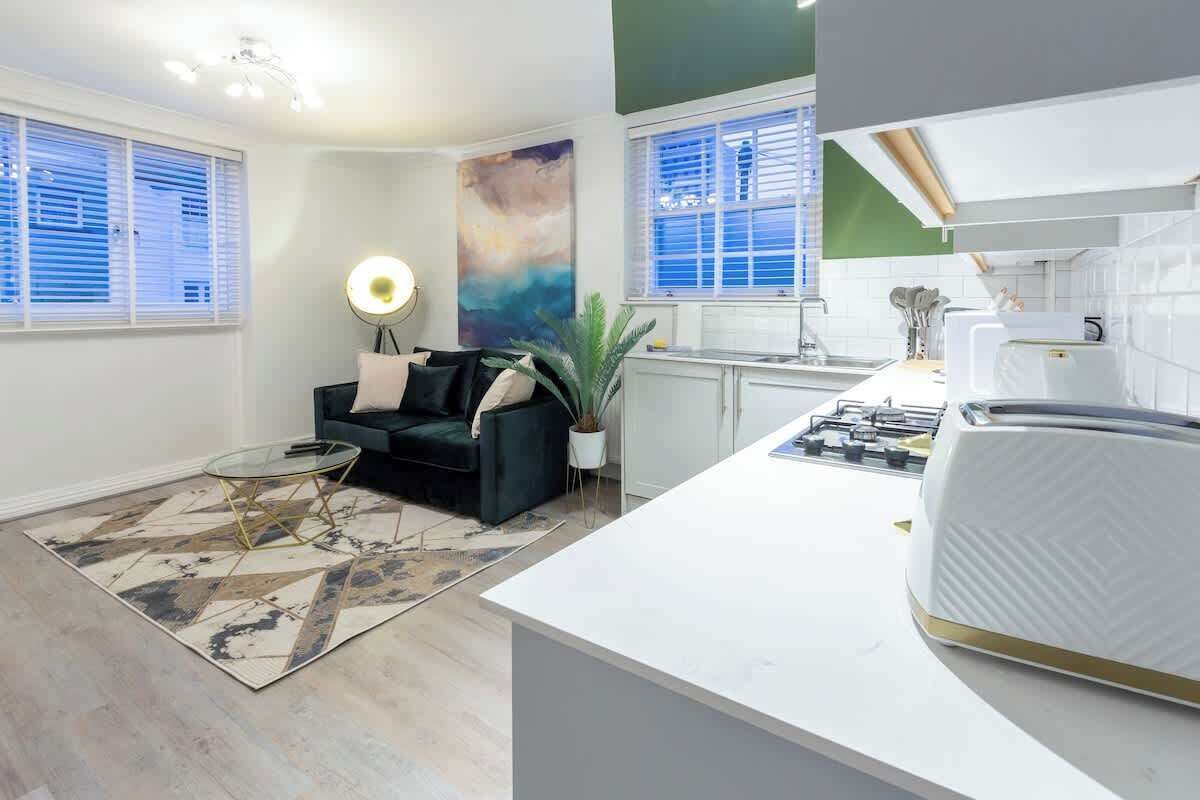 A modern kitchen with white cabinets, a white countertop, and a built-in stove. There is a dining area with a black sofa and a glass coffee table.