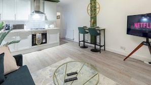 Une cuisine moderne avec des armoires blanches, un four à micro-ondes et une cuisinière à gaz. On y trouve un coin repas avec deux chaises et une table, ainsi qu’un téléviseur fixé au mur qui affiche « NETFLIX ».