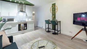 A modern kitchen with white cabinets, a microwave, and a gas stove. There is a dining area with two chairs and a table, and a TV mounted on the wall displaying "NETFLIX".