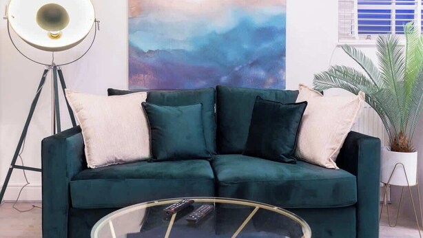 Un canapé vert foncé agrémenté de deux coussins de couleur claire, une table basse en verre avec des télécommandes, un lampadaire et un grand tableau abstrait.