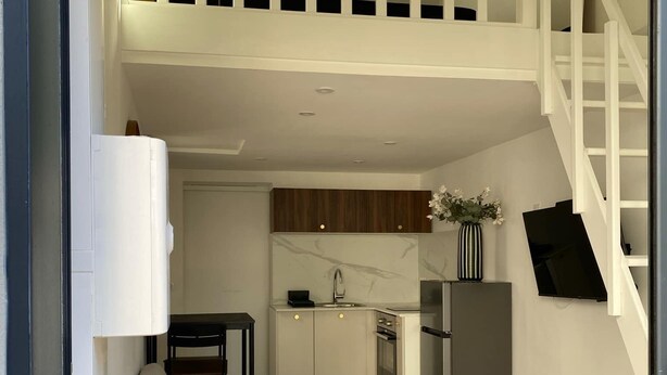 Une cuisine moderne avec des armoires blanches, un comptoir en marbre et un réfrigérateur en acier inoxydable. On y trouve un coin repas avec une chaise et une table, ainsi qu’un escalier menant à l’étage supérieur.