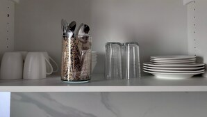 Une étagère de cuisine sur laquelle se trouve un récipient en verre transparent contenant des ustensiles, une pile d’assiettes blanches et quelques tasses blanches.
