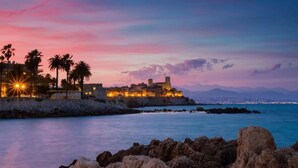 Une vue d’une ville côtière au crépuscule, avec un bâtiment historique fortifié, des palmiers et une mer calme.
