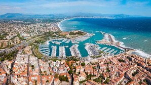 Une vue aérienne d’une ville côtière, avec un port de plaisance rempli de bateaux, entourée de quartiers résidentiels aux bâtiments aux toits rouges.
