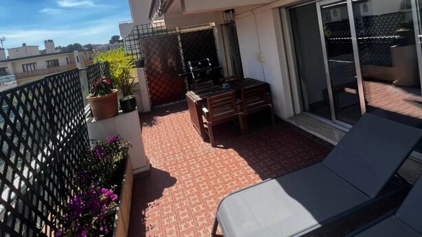Un balcon doté d’un sol à motifs rouges, de deux fauteuils, d’une table et de plantes en pot.
