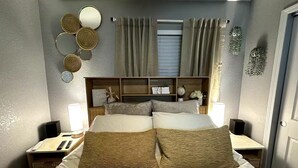 Une chambre bien rangée, avec un lit, deux tables de chevet, une commode, un miroir fixé au mur et une fenêtre avec des rideaux.